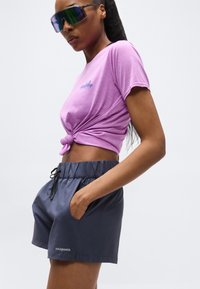 Γυναίκα που φοράει μωβ κοντομάνικη μπλούζα Patagonia με κόμπο, σκούρα σορτς Patagonia και αντανακλαστικά μεγάλα γυαλιά ηλίου μπροστά σε απλό φόντο.