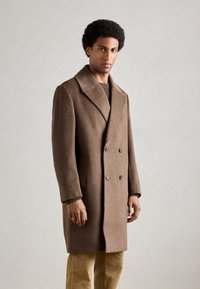 Cappotto doppio petto marrone realizzato in misto lana, con ampie rever e due tasche frontali. Il modello indossa un maglione verde a coste e pantaloni in velluto a costine.