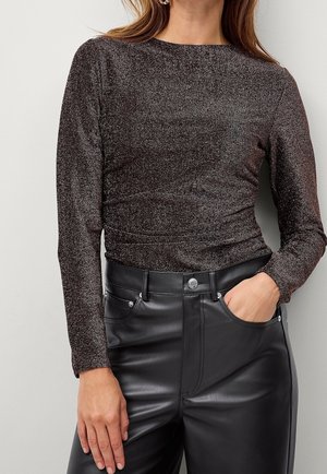 Vrouw die een lange mouw, glinsterende donkerbruine top draagt, in hooggetailleerde zwarte leren broek gestopt, met één hand in de zak.