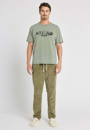 Homme debout portant un t-shirt à manches courtes vert olive avec un motif de poisson, un pantalon en velours côtelé olive avec cordon de serrage, et des chaussures beige faciles à enfiler.