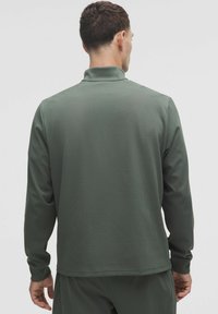 Tmavozelený dlhý rukáv pulóver vyrobený z textúrovanej látky, s stojatým golierom a uvoľneným strihom, bezšvíkové ramená.