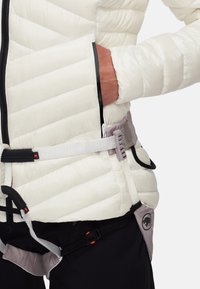 Giacca bianca trapuntata con cerniera nera e strap elastici. Presenta una vita grigia con una tasca laterale e accenti neri sui pantaloni.