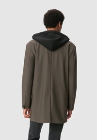 Hombre con un abrigo largo marrón y capucha negra, de pie de espaldas contra un fondo gris claro liso.