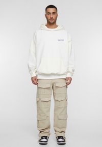 Hvid hættetrøje med en frontlomme og lilla grafik. Sammen med beige cargo bukser med flere lommer. Sorte sneakers.