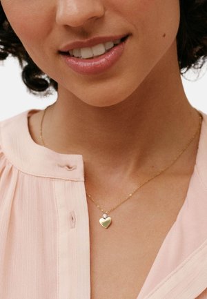 Ogrlica z obeskom v obliki zlate srčke z majhnim kamnom, nošena čez mehko roza gubano bluzo, ki poudarja nežno zasnovo verižice.