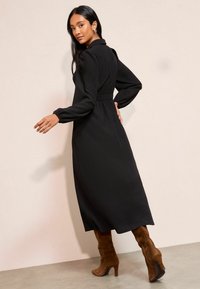 Černé dlouhé šaty s límcem, elastickým pasem a volnou sukní. Modelka má na sobě hnědé semišové kozačky po kolena. Hladká textura látky.