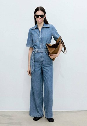 Jeansoverall med korta ärmar, midjebälte och vida ben; har en bröstficka och sliten blå färg. Modellen håller en brun väska.