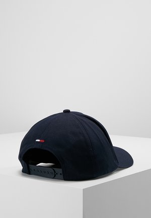 Czarna regulowana czapka typu snapback z małym czerwono-biało-niebieskim detalem flagi z tyłu, umieszczona na białej powierzchni na tle szarego tła.