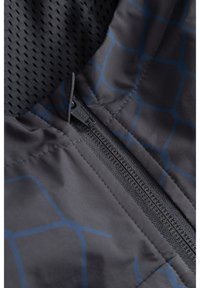 Közeli kép egy fekete cipzárról sötét anyagon, enyhe kék kockás mintával és lyuggatott fekete béléssel alatta.