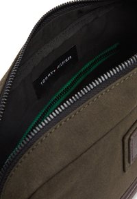 Olivengrønn glidelåseveske med svart stofffôr, med en Tommy Hilfiger-tag og en grønn glidelåslomme. Teksturet ytre overflate.