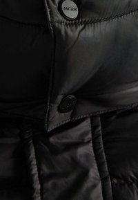 Veste matelassée noire avec un extérieur en nylon lisse et brillant, un motif matelassé proéminent, et des boutons-pression noirs avec logo.