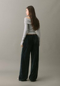 Nainen, jolla on pitkä ruskea hius, pukeutuneena harmaaseen olkaimeton paita ja leveät mustasta sametista housut, seisoo yksinkertaista harmaata taustaa vasten.