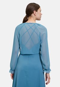 Robe bleu clair avec des manches longues transparentes, présentant un dos court au design croisé. Texture douce et légers volants aux poignets.