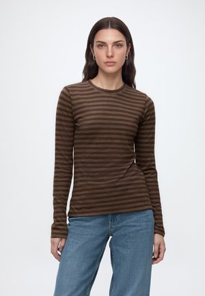 Young woman with long dark hair wearing brown striped long-sleeve shirt and blue jeans, standing against a plain white background.