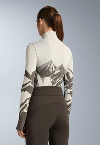 Priliehavý top s dlhým rukávom v svetlosivej farbe so vzorom hôr v tmavosivej farbe. Hladká textúra, vysoký golier a kombinovaný s tmavohnedými legínami.