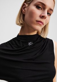 Melnā bez piedurknēm topiņa ar savilkta auduma detaļām un augstu kakla izgriezumu. Uz priekšpuses ir balts izšūts logo, patīkama gluda tekstūra un pieguļošs dizains.