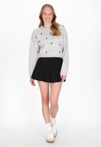 Jasnoszary sweter z wielokolorowymi wzorami w kształcie serc, zestawiony z czarną plisowaną spódnicą. Białe trampki i skarpetki dopełniają outfit.