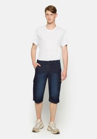 Shorts cargo en denim foncé avec deux poches latérales et une ceinture épurée, associés à un t-shirt blanc ajusté et des baskets neutres.