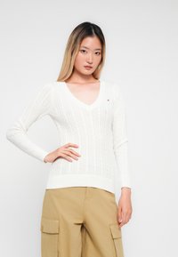 Mulher jovem com cabelo longo, vestindo um suéter branco de malha em V e calças cargo bege, de pé com uma mão na anca.