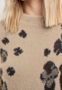 Pull beige avec des taches noires de motif animal, présentant une texture douce et duveteuse et un col rond. Gros plan sur les détails du tissu et du motif.