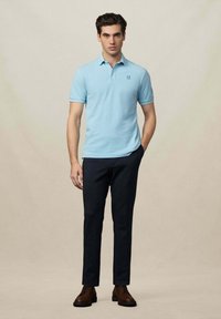 Polo shirt azzurro chiaro con colletto e logo ricamato, abbinato a pantaloni scuri su misura e scarpe stringate marroni.