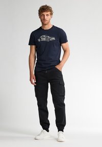 Námořnická modrá bavlněná tričko s grafikou auta ve světlé barvě, kombinované s černými cargo kalhotami s bočními kapsami a kotníkovými manžetami, doplněné o bílé tenisky.