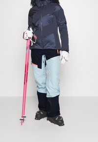 Skidkläder inklusive en marinblå quiltad jacka med pälsluva, ljusblå skidbyxor med mörka accenter, och rosa skidstavar.