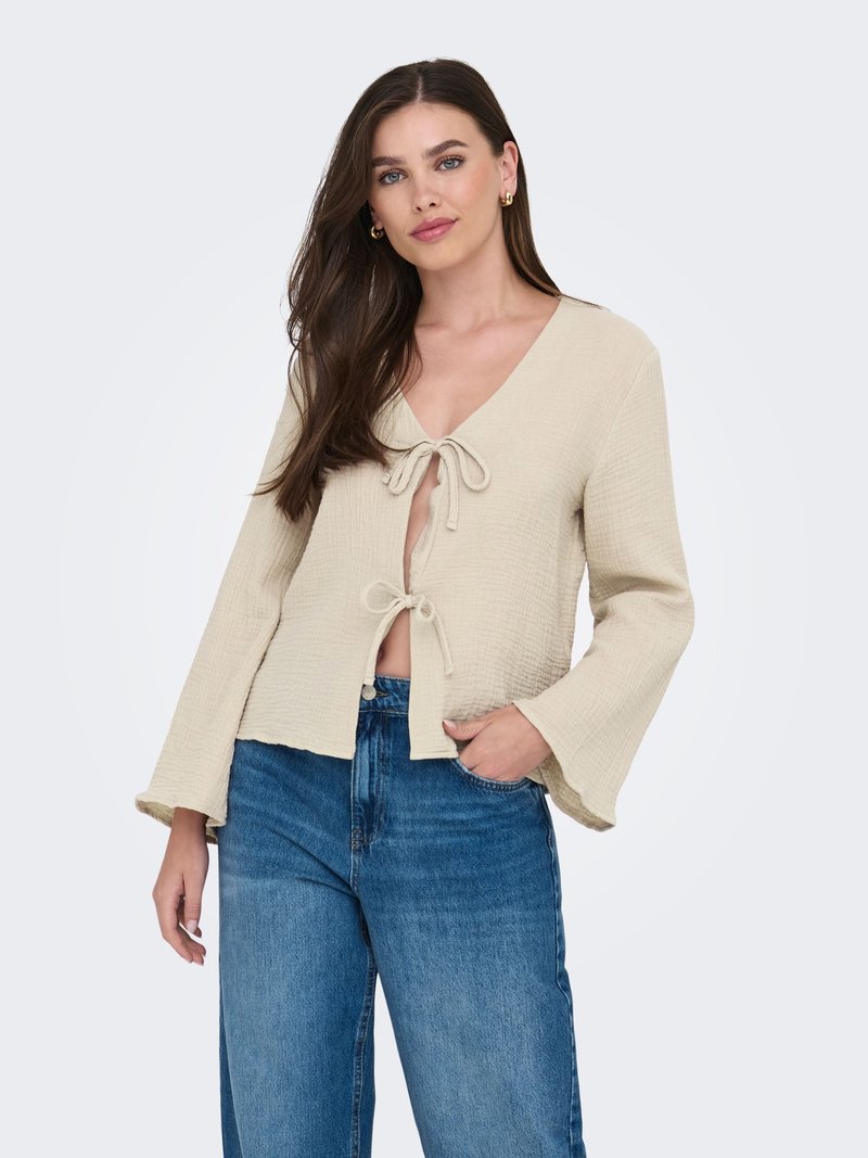 Femme aux longs cheveux bruns portant un haut beige texturé noué avec deux nœuds et un jean bleu à jambes larges, debout avec une main dans la poche.