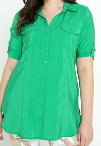 Donna che indossa una camicia verde brillante con bottoni, due tasche sul petto e maniche arrotolate, abbinata a pantaloni leggeri con motivo floreale.