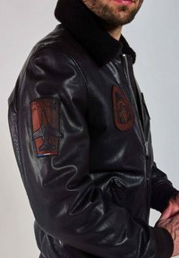 Homme portant une veste en cuir noire avec un col en fourrure noire, des écussons sur le bras et la poitrine, y compris un avion et le texte "Patrouille de France".