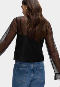 Blusa negra transparente con un acabado texturizado, mangas largas transparentes y una capa interior negra ajustada. Combinada con jeans de mezclilla azules.