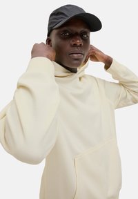 Pullover hoodie i blødt cremefarvet stof med høj krave, kængurulomme og afslappet pasform. Sort kasket komplementerer outfittet.