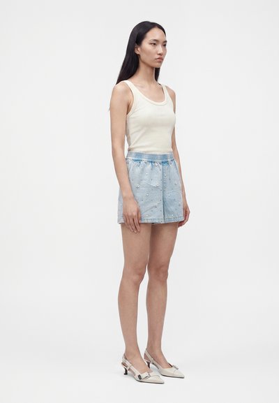 Jeune femme debout portant un haut blanc sans manches, un short bleu clair brodé et des chaussures blanches à talons bas et bride arrière sur un fond uni.