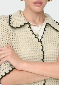 Béžový pletený cardigan s černými detaily, s vroubkovaným límcem, pěti knoflíky a otevřenou texturou. Akcenty zlatého a perlového šperku.
