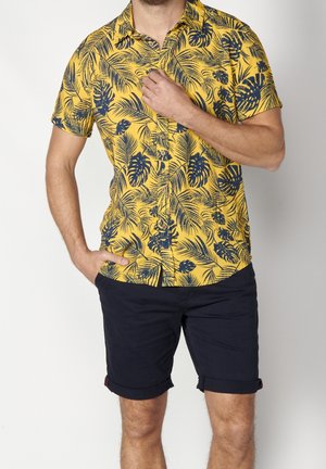 Uomo che indossa una camicia gialla a maniche corte con motivi di foglie blu scuro e pantaloncini blu scuro risvoltati, mano che sistema il colletto della camicia.