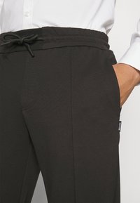 Schwarze Jogginghosen mit elastischem Bund, Kordelzug, seitlichen Taschen und einer dezenten Textur. Mit einem kleinen Etikett an der Seite.