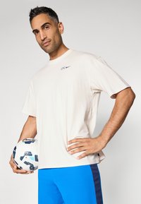 Kremowa koszulka z okrągłym dekoltem z napisem "CFC" oraz logo Nike w kolorze fioletowym, zestawiona z jednolitymi, niebieskimi szortami sportowymi. Trzyma w dłoniach wzorzystą piłkę nożną.