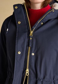 Primo piano di una persona che indossa una giacca blu navy con zip e bottoni dorati sopra un maglione rosso a coste.