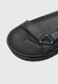 Sorte lærsandaler med en struktureret overflade, der har to brede stropper og en gummisål. Inkluderer hardware i sølvfarve, hvor stropperne forbindes.