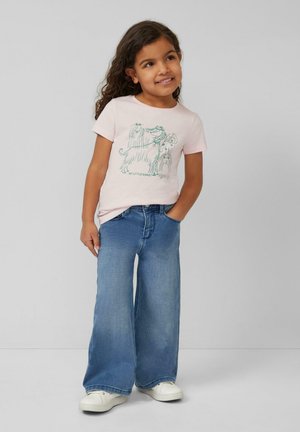 Junges Mädchen mit lockigem Haar, das ein rosa T-Shirt mit Hundemotiv, weite blaue Jeans und weiße Turnschuhe trägt und mit einer Hand in der Tasche steht.