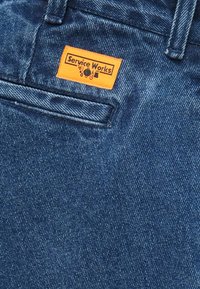 Sinised teksapüksid, millel on tagataskus oranž "Service Works" silt, millel on puhastuse ja tööriistade ikoonidega logo.