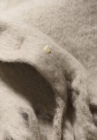 Beige Wollschal mit einer weichen, flauschigen Textur, ausgestattet mit einem dekorativen beigen Knopf und fransigen Kanten.