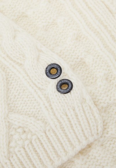 Cremefarvet strikket sweater med kabelmønster, ledsaget af to sorte knapper med indgraveret tekst og en cirkulær metalaccent.