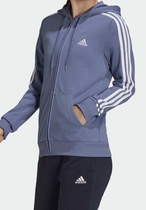 Osoba ubrana w niebieską bluzę z kapturem i zamkiem na całej długości marki Adidas, z białymi paskami na rękawach, oraz czarne spodnie Adidas z logo na udzie.
