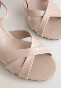 Paire de sandales en cuir verni beige avec des lanières croisées et des orteils ouverts sur une surface claire.