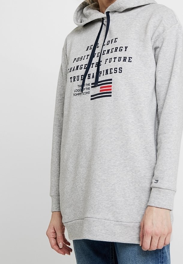 Hoodie gris clair avec cordon de serrage, comportant un texte et un logo de drapeau sur le devant. Le tissu a une texture douce et une coupe décontractée.