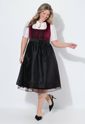 Mujer sonriente con vestido tradicional con blusa blanca de encaje, corpiño burdeos, delantal negro atado a la cintura y zapatos negros de tacón con lazos.
