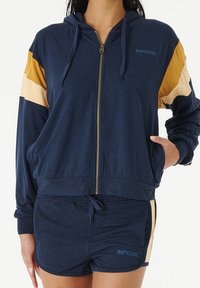 Sweat à capuche zippé bleu marine avec des accents rayés or et crème, doté d'une capuche à cordon et de poches avant. Accompagné d'un short assorti.