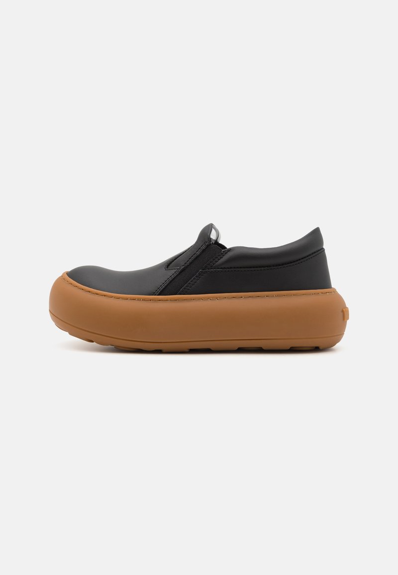 Zapatos negros sin cordones con punta redonda, hechos de cuero suave, con una suela de goma marrón claro y una pestaña en la parte trasera para facilitar el calzado.