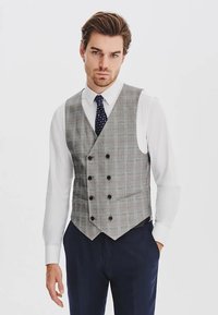 Mężczyzna w białej koszuli, granatowym krawacie w wzory, szarym dwurzędowym kamizelce w kratę i granatowych spodniach, stojący z jedną ręką w kieszeni.